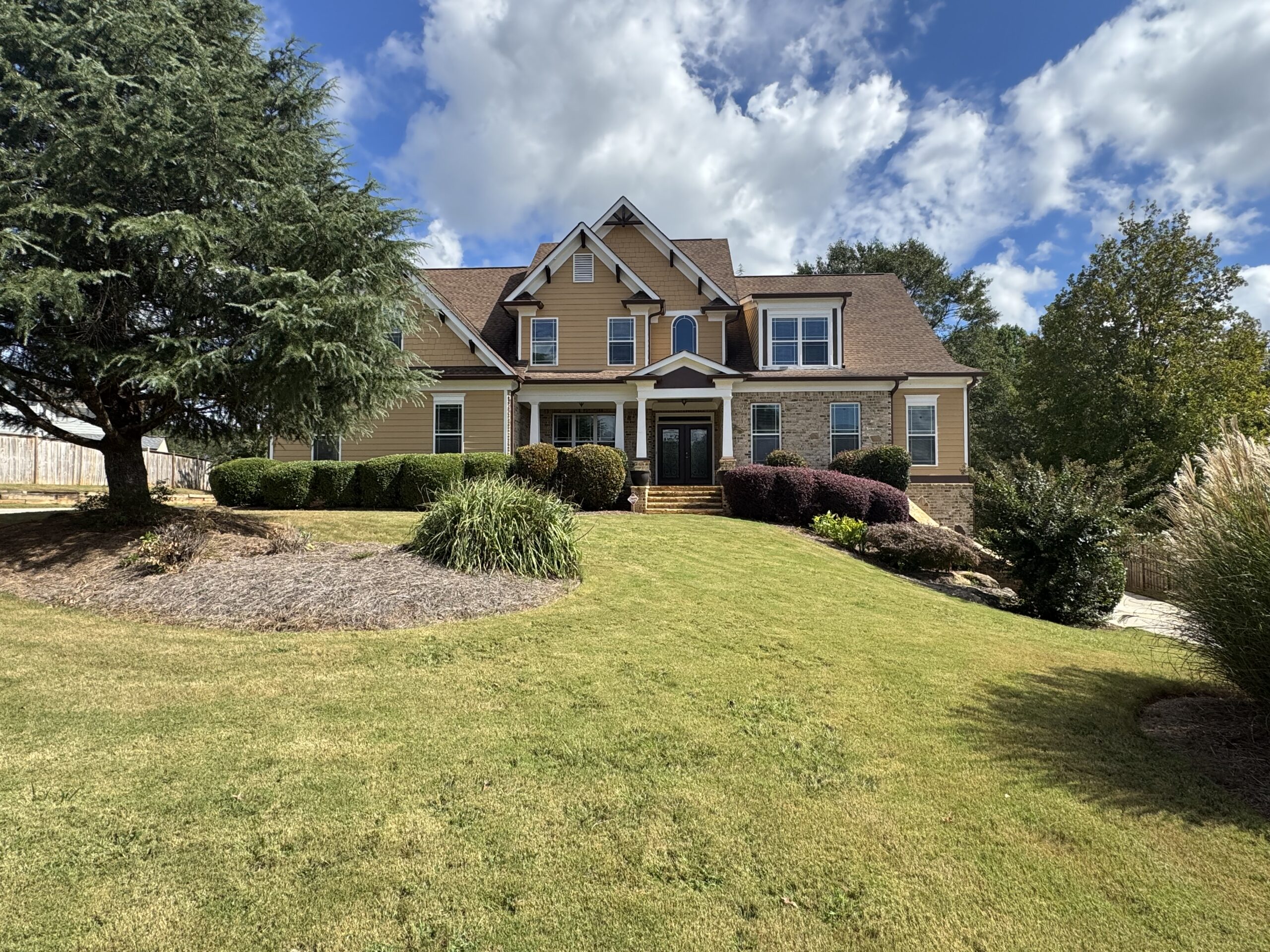 A large two-story house with tan siding, dark shutters, and a landscaped front yard with bushes and trees under a partly cloudy sky.