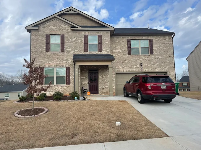 Two-story brick house with brown shutters, a single-car garage, and a red SUV parked in the driveway on a cloudy day. The lawn is yellowish and there is a small tree in front.