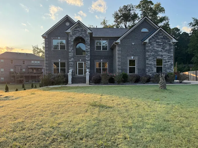 Two-story brick house with multiple windows and a large front lawn, photographed in the early morning or late afternoon sunlight.