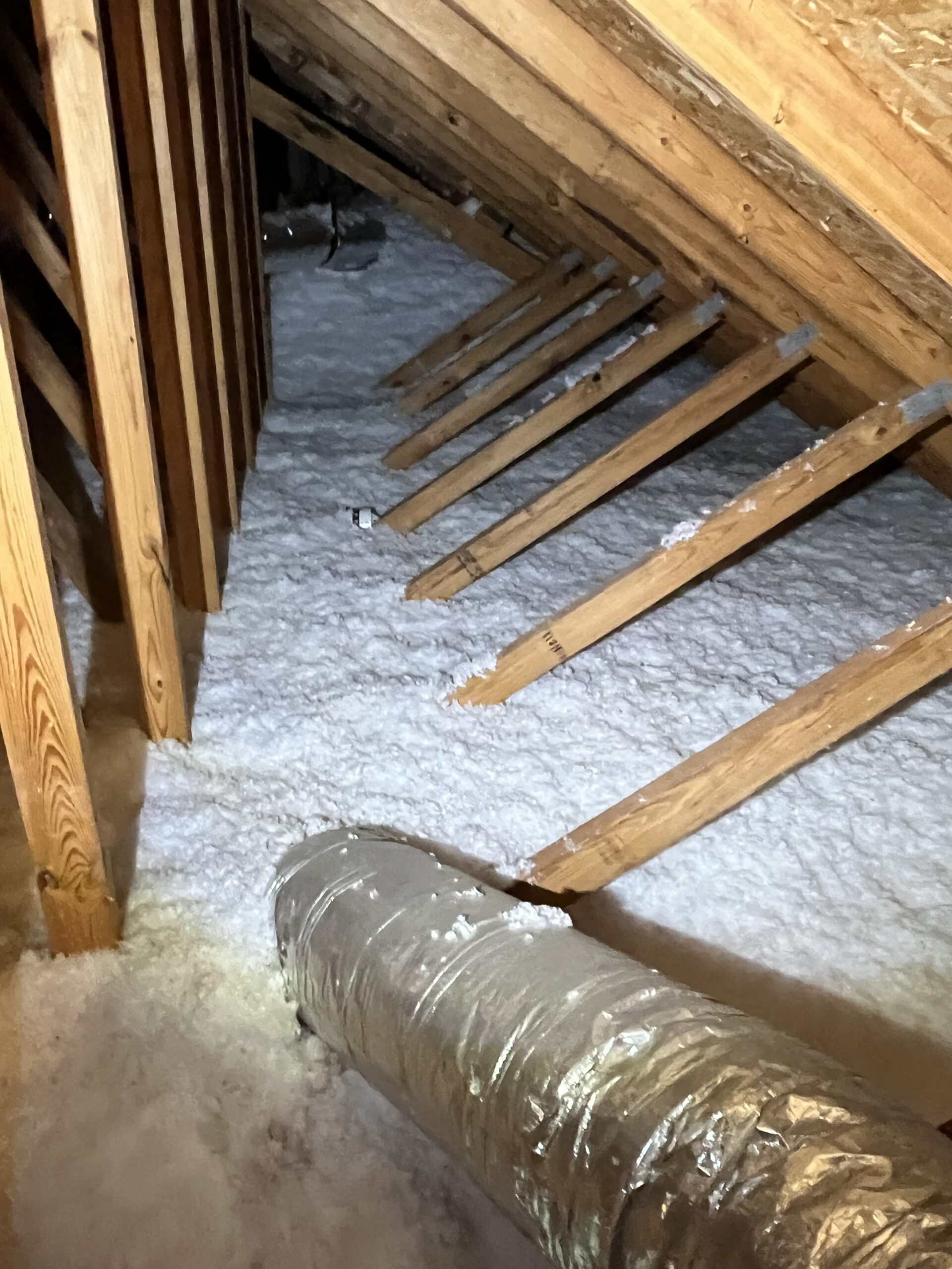 Attic with wooden rafters, blown-in insulation covering the floor, and a large metal air duct running along the bottom. The area is illuminated by artificial light.
