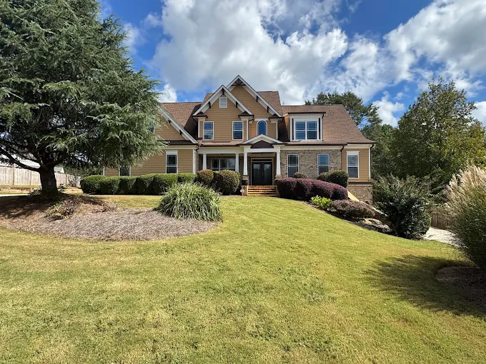 A two-story yellow house with white trim sits on a sloped lawn, surrounded by manicured shrubs, trees, and a wooden fence under a partly cloudy sky.