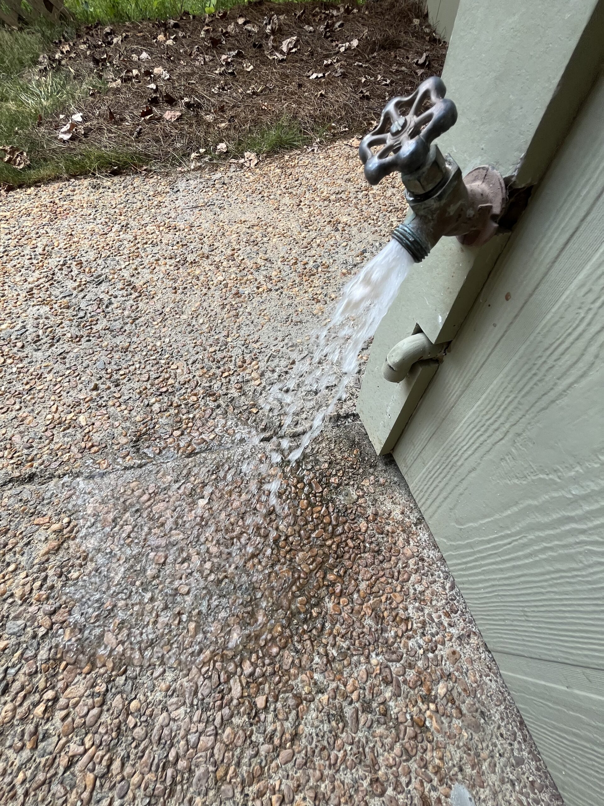 Water flows from an outdoor faucet attached to a light green wall, splashing onto a textured stone ground.