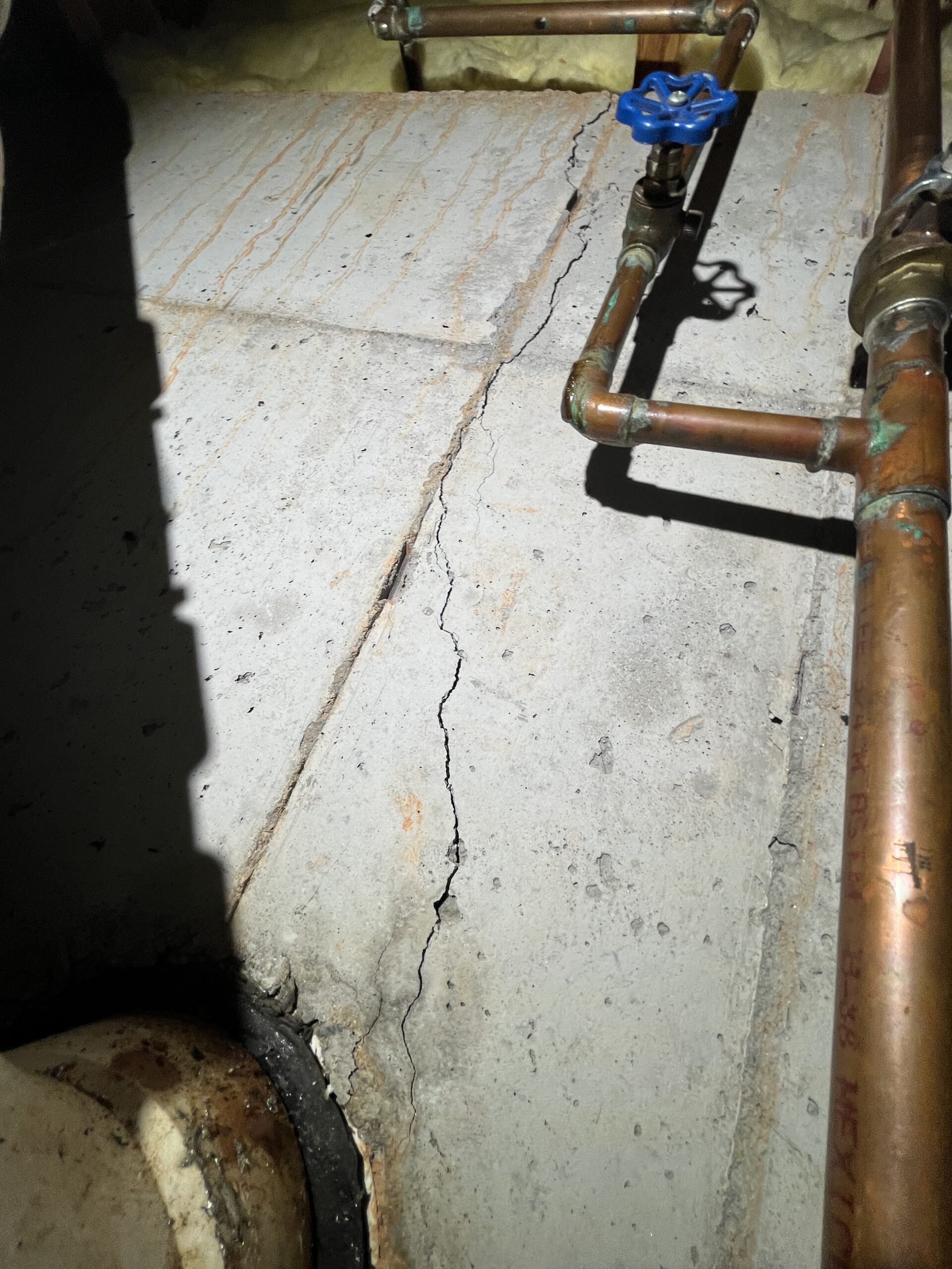 A visible crack runs along a concrete floor next to copper pipes and a blue valve under artificial light.